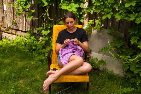 A young woman in a black T-shirt and jeans shorts knits a sweater from natural lilac woolen threads and sits on an armchair in the country on the background of a green lawn