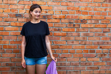 A young woman in a black T-shirt and jeans shorts stands near a red brick wall and holds in her hands knitting needles and an untied sweater made from natural lilac wool threads at the cottage