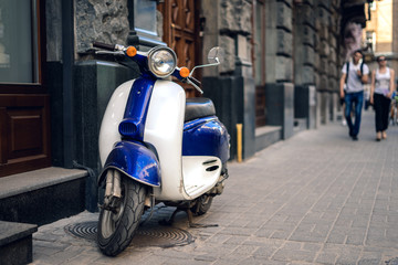 Fototapeta na wymiar Blue scooter on background of old city. Scooter parked on sidewalk of empty city street.