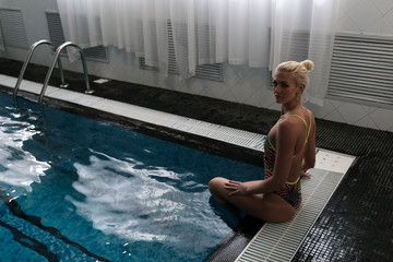 An attractive blonde sits on the side of the pool and looks into the frame. Photo above.