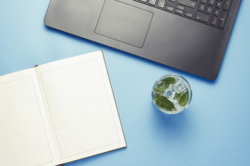 Workplace with Laptop, a fresh drink with ice and mint, an open diary on a blue background. The...