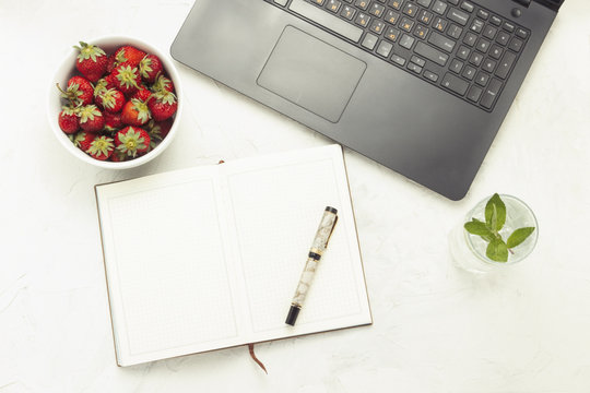 Laptop, diary, fresh drink with ice and mint and a white plate with strawberries on a light stone background. Concept of working at home in a cozy atmosphere. Flat lay, Top view