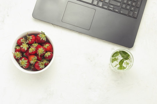 Laptop, fresh drink with ice and mint and a white plate with strawberries on a light stone background. Concept of working at home in a cozy atmosphere. Flat lay, Top view