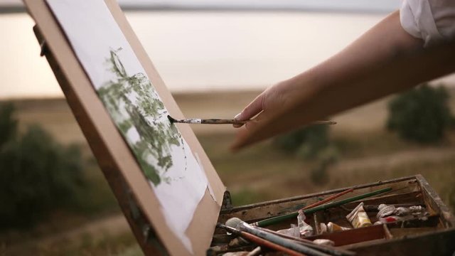 Close up of artist working on her future picture standing outdoors on a green meadow near the lake. Puts colours on canvas using a paintbrush and palette