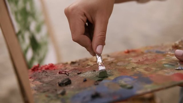 Close up of a artist female's hand mixing paints on palette and applies it on her surface on easel. Working process. Outdoors