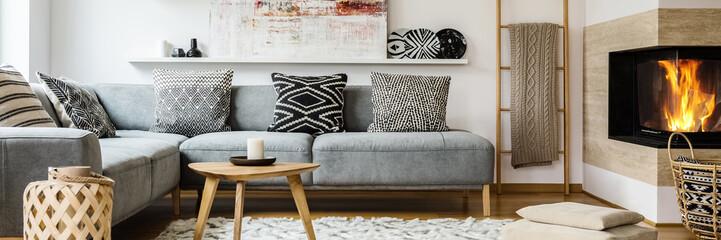 Real photo of modern painting placed on the shelf above grey sofa with patterned pillows standing in bright sitting room interior with blanket on wooden ladder and fireplace