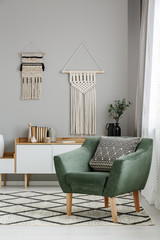 Real photo of a boho living room interior with macrame hanging on gray wall behind a comfy, green...