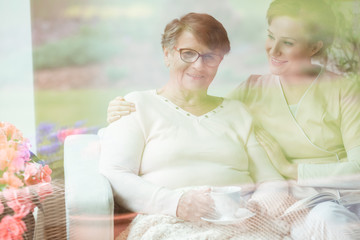 Senior woman drinking tea, sitting on a couch in a day care center with her caregiver. Behind the...