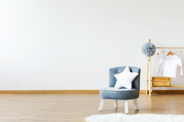 Star shaped white pillow placed on small armchair standing in white baby room interior with grey pompom and white shirt on wooden hanger. Empty wall for your poster - obrazy, fototapety, plakaty