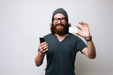 Cheerful smiling bearded hipster man holding smartphone and credit card, online shopping concept