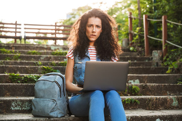 Photo of upset confused woman sitting on stairs in park on summer day, and using silver laptop with disappointment