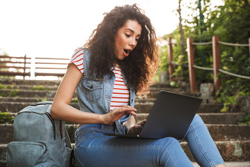 Photo of excited brunette woman sitting on stairs in park on summer day, and using silver laptop with surprise