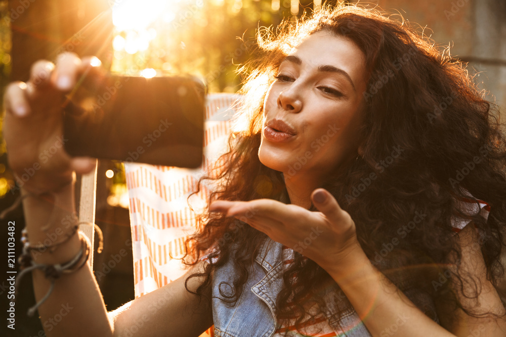 Poster photo of seductive flirty woman 18-20 smiling and blowing air kiss at cell phone, while taking selfi