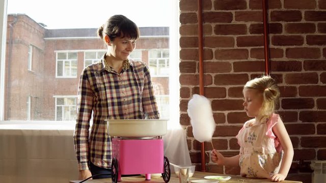 A young mother makes cotton candy on a special machine, a little girl watches the process and waits for a dessert indoors with brick walls.