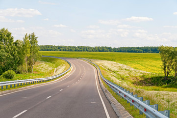 Naklejka premium Steep highway turn with metal rails along against blossoming meadow and forest background. Kaluzhsky region, Russia. 