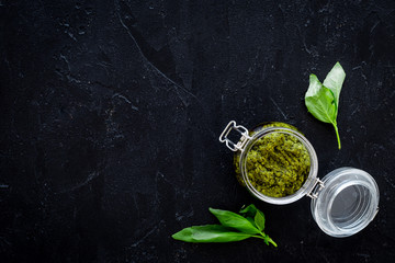 Green pesto sauce in glass jar near basil leaves on black background top view copy space