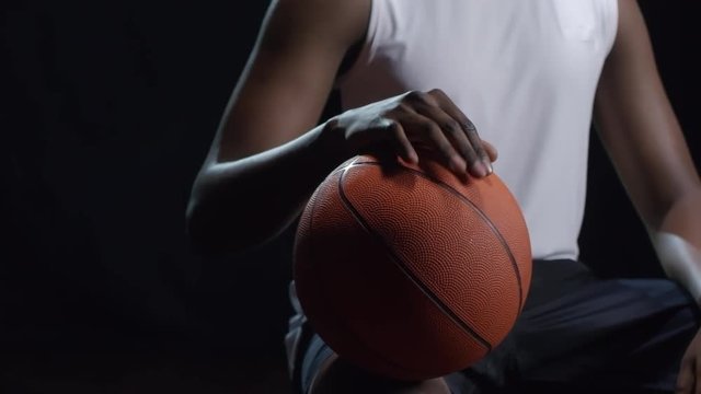 Studio shot with PAN of unrecognizable black basketball player sitting isolated on dark background and tapping fingers on ball