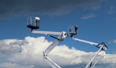 Two white cherry pickers against blue sky with clouds, Under there are fluffy clouds, above dark clouds