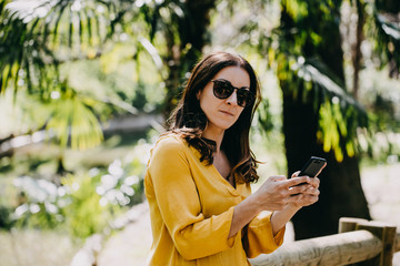 .Pretty young woman wearing a brown hat walking around a famous park in Madrid in a summer day using her mobile phone, talking and texting. Lifestyle portrait.