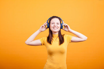 Beautiful young woman listening music in studio from her headphones on yellow background