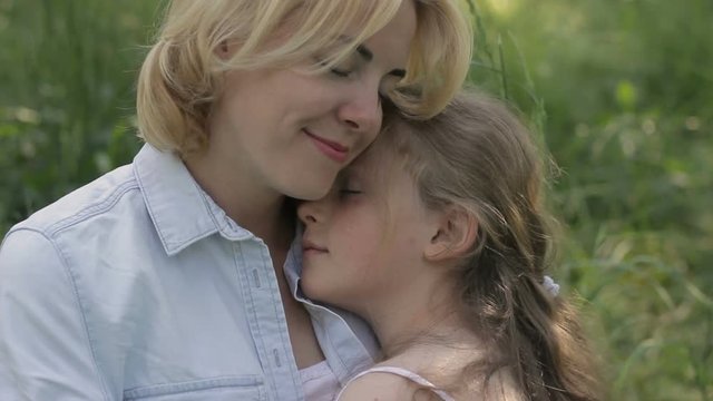 Young mother and daughter with close eyes sitting in park, embracing, stroke