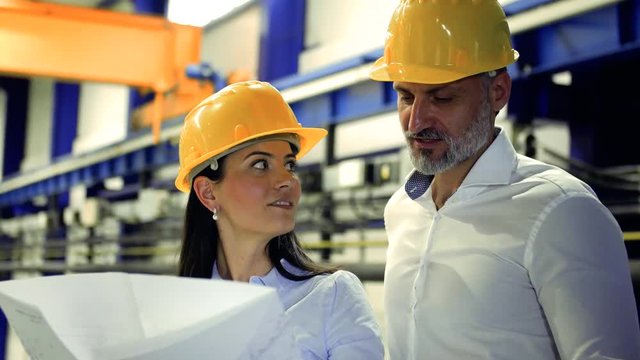 Industrial man and woman engineers with yellow helmets looking at blueprints.