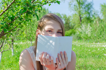 Young pretty woman in white top with long hair sitting under apple tree and hides face behind book on sunny day.