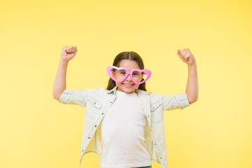 Loves power. Kid girl heart shaped eyeglasses cheerful posing with power gesture. Girl curly hairstyle adorable smiling face. Child charming smile yellow background. Kid happy lovely enjoy childhood