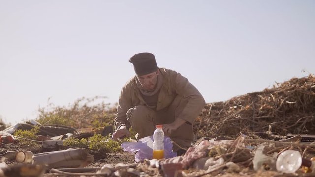 dirty homeless hungry man in a dump eating orange for food in lifestyle the package with walking goes looking for food slow motion video. homeless dirty man roofless person looking for food in a dump