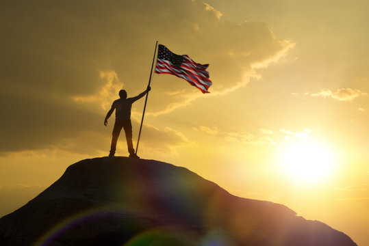 The symbol of a man with the flag of the United States of America stands on the top of the mountain. Against the sky and the sun at sunset. Success and achievements, patriotism. Independence Day. USA