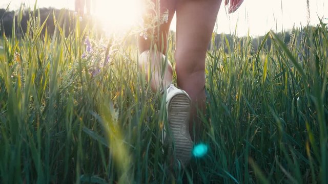 Young beautiful woman in sneakers and shorts walking on meadow with green grass nature slow motion video. Girl in the field legs at sunset close-up on the grass sunlight lifestyle silhouette
