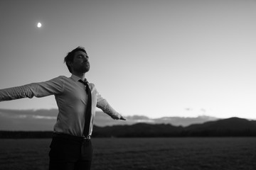 Greyscale image of businessman in white shirt and black tie standing outdoors with open arms