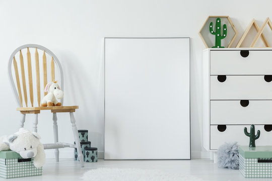 Fluffy toy placed on wooden chair in white baby room interior with cupboard with decor, boxes and mockup poster. Paste your graphic here