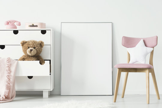 Empty poster with place for your graphic standing on the floor in white room interior with pink wooden chair and teddy bear placed in cupboard's shelf