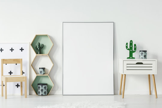 Small cupboard with cactus shaped lamp and decorative box standing in white baby room interior with empty poster with place for your graphic, wooden chair and geometric shelves