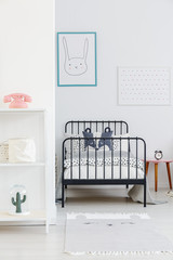 Child's bed with black metal frame in a small, simple bedroom interior. Rabbit poster on a white wall. Real photo