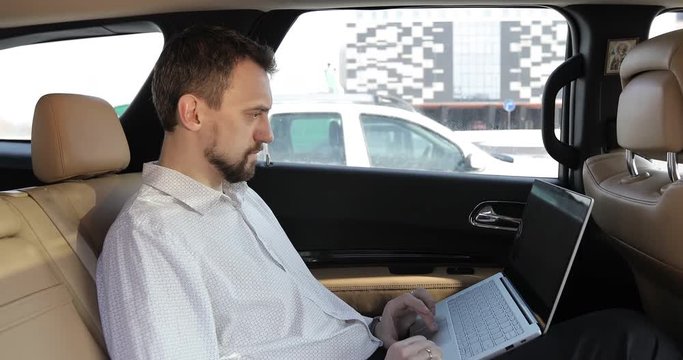 Businessman with a beard working on a laptop on backseat of a car.