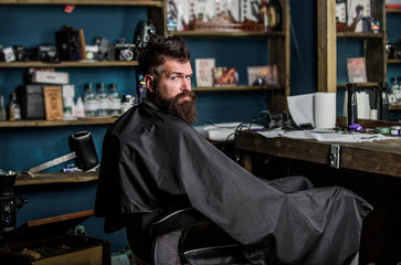 Man with beard covered with black cape sits in hairdressers chair in front of mirror. Hipster with beard waits for barber and haircut. Man with beard client of hipster barbershop. Barbershop concept