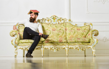 Macho sits with open book on head, like roof. Man with beard and mustache sits on sofa, white wall...