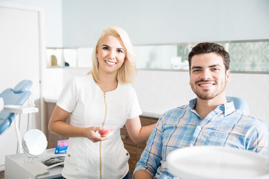Beautiful dentist designing a smile and holding a teeth model next to a patient in a dental clinic