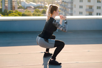 Side view of young sporty woman in stylish sportswear doing squat and bob exercise during the workout on the building rooftop on sunny day.
