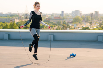 Attractive blonde fit and sporty woman in stylish sportswear doing the jump rope exercise during the workout on the building rooftop on sunset.