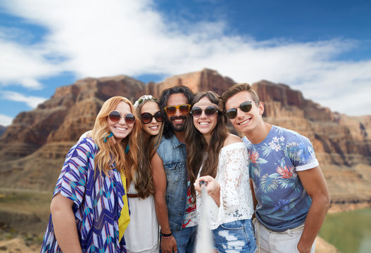 travel, tourism and technology concept - smiling young hippie friends taking picture by smartphone on selfie stick over rocks of grand canyon national park background