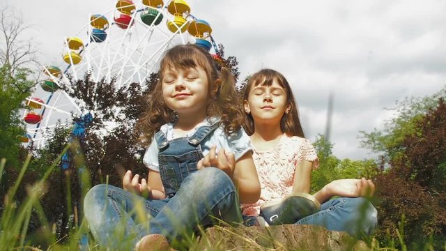 The children sit on the grass in a lotus pose. Sisters are meditating in the park.