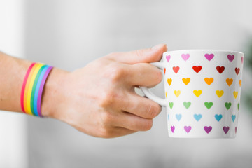 homosexual and lgbt concept - close up of female hand with cacao drink in cup with heart pattern and gay pride awareness wristband on wooden table