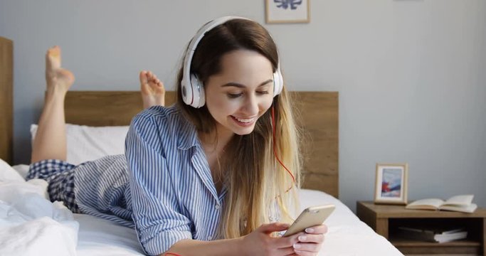 Young beautiful Caucasian woman in the headphones lying on her stomach on the unmade bed in the pajama and listening to the music on the smartphone. Indoors