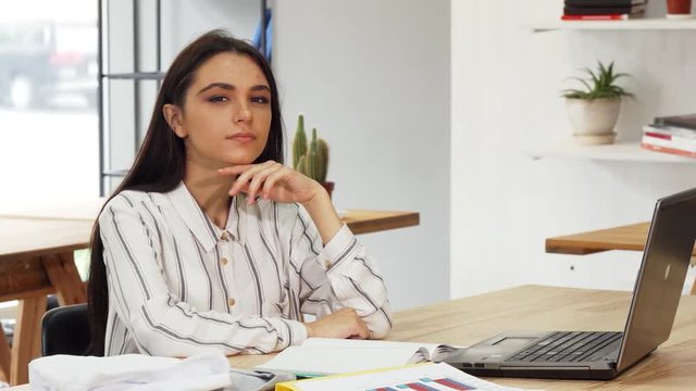 Portrait of a young beautiful businesswoman sitting in front of her laptop in a modern office. Attractive female designer looking thoughtfully to the camera, while working on a computer.
