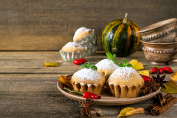 Autumn still life. Homemade cupcakes with powdered sugar with cinnamon sticks, anise stars, pumpkins, berries of rosehip and autumn leaves on rustic wooden background.