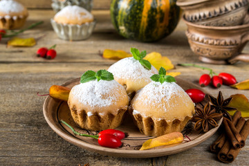 Autumn still life. Homemade cupcakes with powdered sugar with cinnamon sticks, anise stars, pumpkins, berries of rosehip and autumn leaves on rustic wooden background.