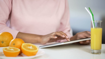 Mixed race lady using tablet, oranges and juice on table, health article search